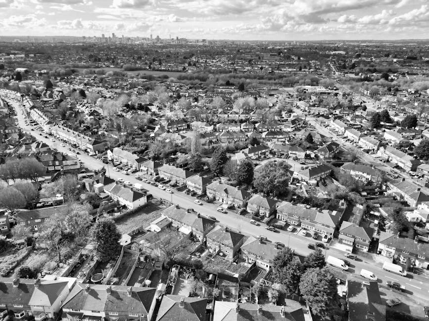 Aerial black-and-white view of a residential neighbourhood showing rows of terraced and semi-detached houses with tiled roofs, front gardens, and parked cars lining the streets. The scene captures a w